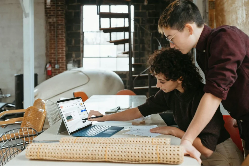Two colleagues collaborating at a desk in a rustic loft office, looking together at a laptop screen.