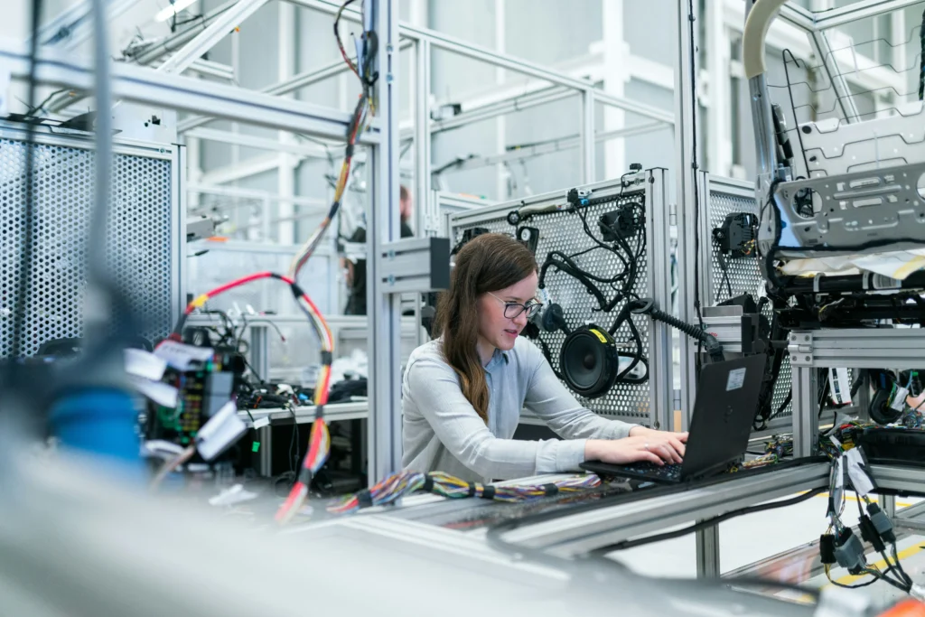 A woman works intently on a laptop amidst a high-tech engineering lab, surrounded by complex machinery and colorful wiring, conveying innovation.