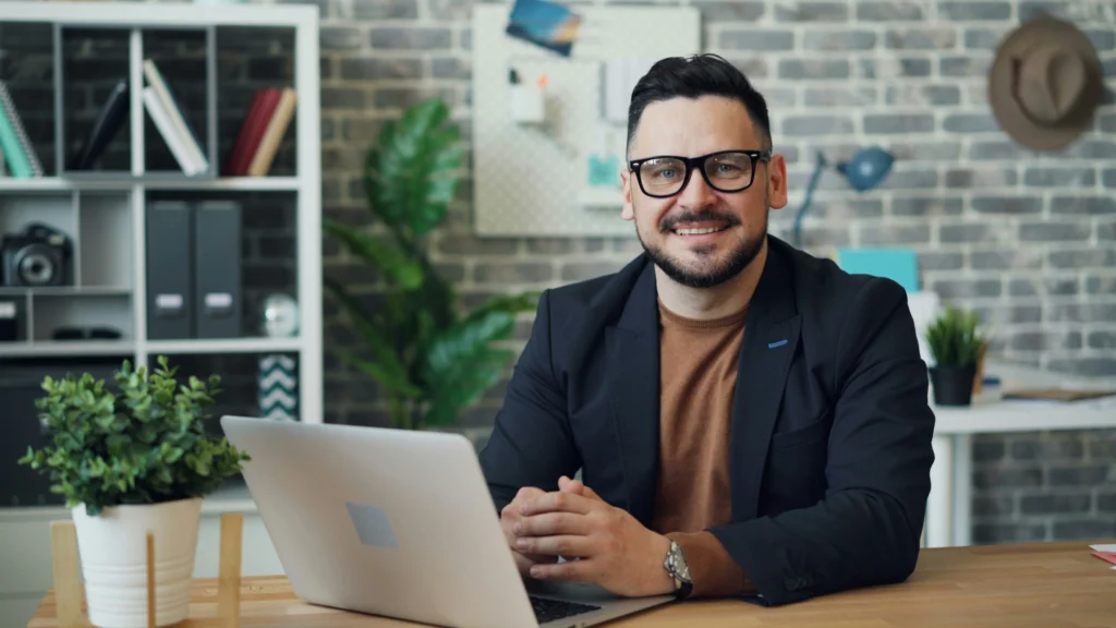 Smiling man with glasses and a beard sits at a desk with a laptop, in a modern office. Shelves, plants, and a brick wall create a cozy atmosphere.