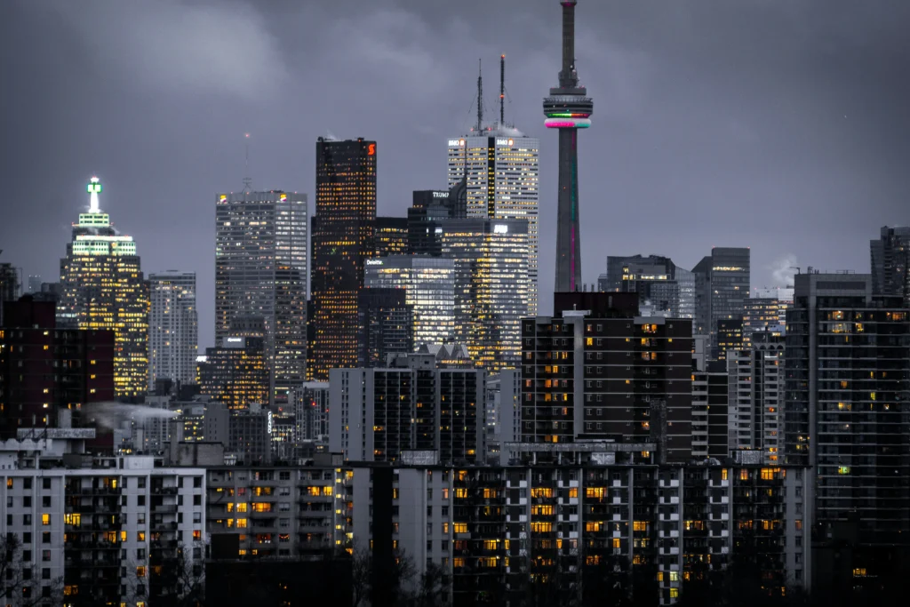 City skyline at dusk with illuminated buildings and the CN Tower. The overcast sky adds a dramatic tone to the bustling urban scene.