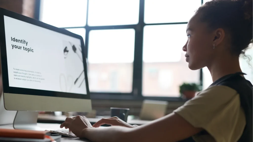 Here is a descriptive alt text for the image: Alt Text: A side-profile view of a young woman with her hair tied up in a bun, focused on a large desktop computer monitor in a bright office setting. Her hands are placed on the keyboard as she types. The computer screen displays a presentation slide that reads "Identify your topic" in large, bold text, followed by a smaller paragraph underneath. In the background, a large grid window lets in soft, natural light, showing a blurred view of another desk and a small potted plant.