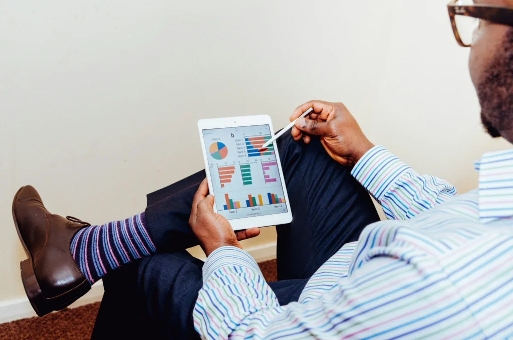 A man wearing a striped shirt and patterned socks sits with legs crossed, pointing a white stylus at business charts on a digital tablet.