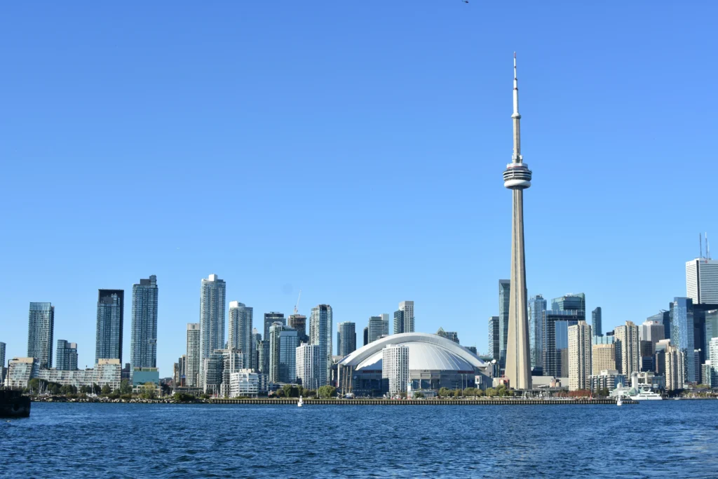 Panoramic view across blue water toward the iconic Toronto city skyline, featuring the CN Tower and Rogers Centre under a clear blue sky.