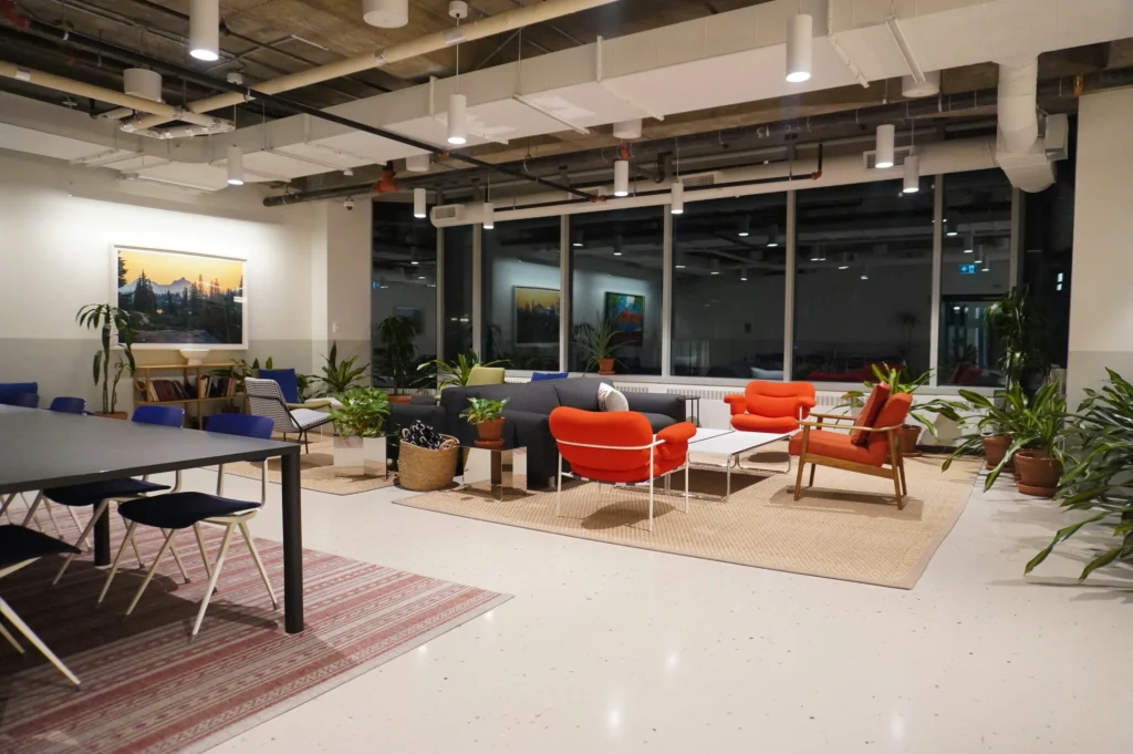 Modern office lounge area featuring orange armchairs, a gray sofa, house plants, and a conference table near floor-to-ceiling windows.