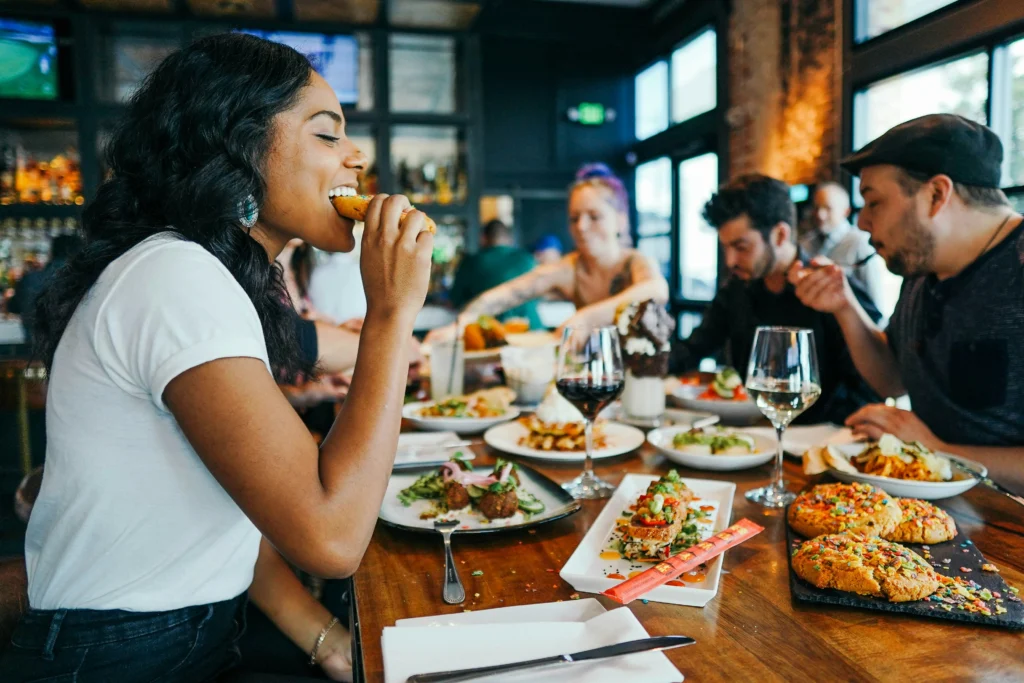Diverse group of friends enjoys food and drinks together around a dining table at a lively restaurant during a festive social gathering.