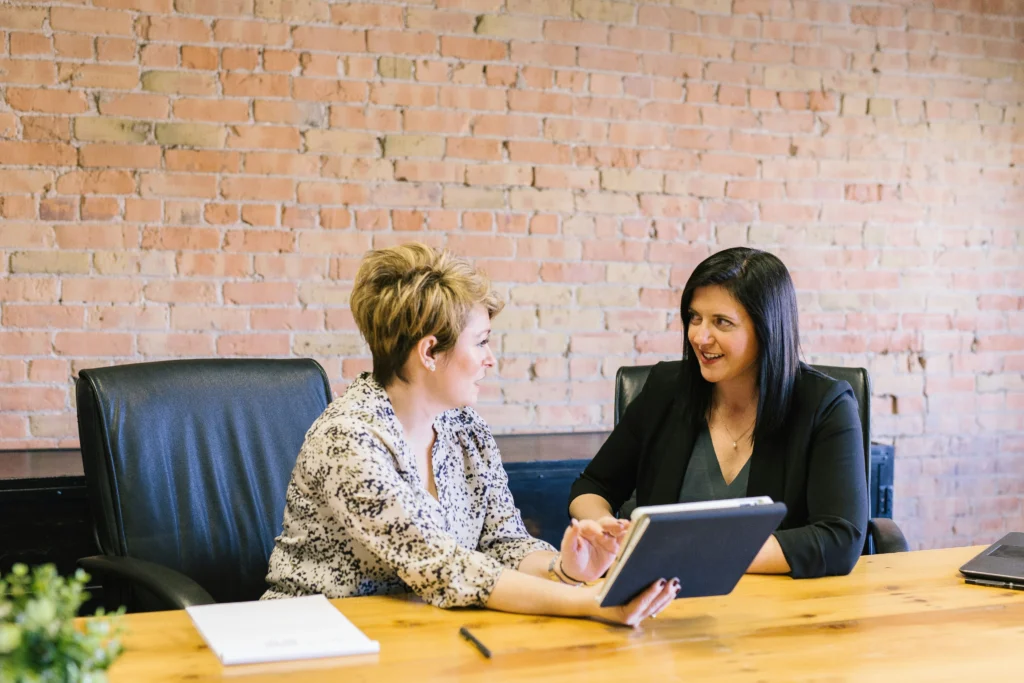 Two professional women sit at a wooden desk in front of an exposed brick wall, discussing information shown on a tablet screen.