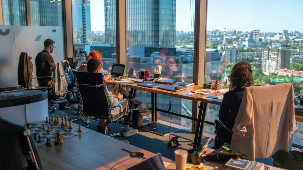 Employees work at laptops along a long window desk inside a high-rise office offering expansive views of the surrounding city skyline.