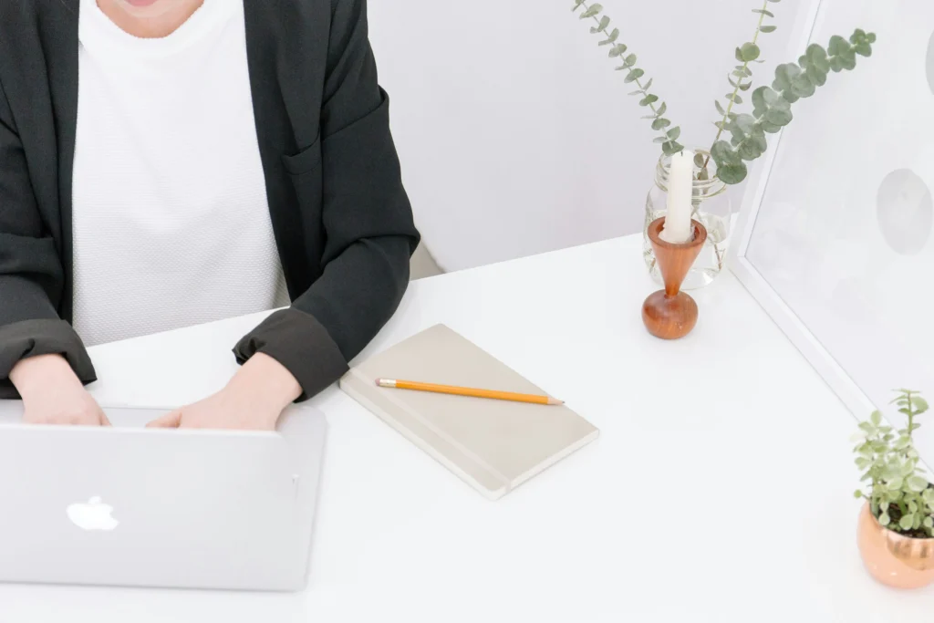 Overhead view of a person in a black blazer typing on a laptop at a bright white desk with a notepad, candle, and potted plants.