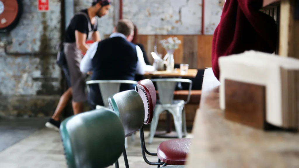 Retro green and red vinyl chairs sit in the foreground of a rustic, industrial restaurant where a waiter serves customers in the background.