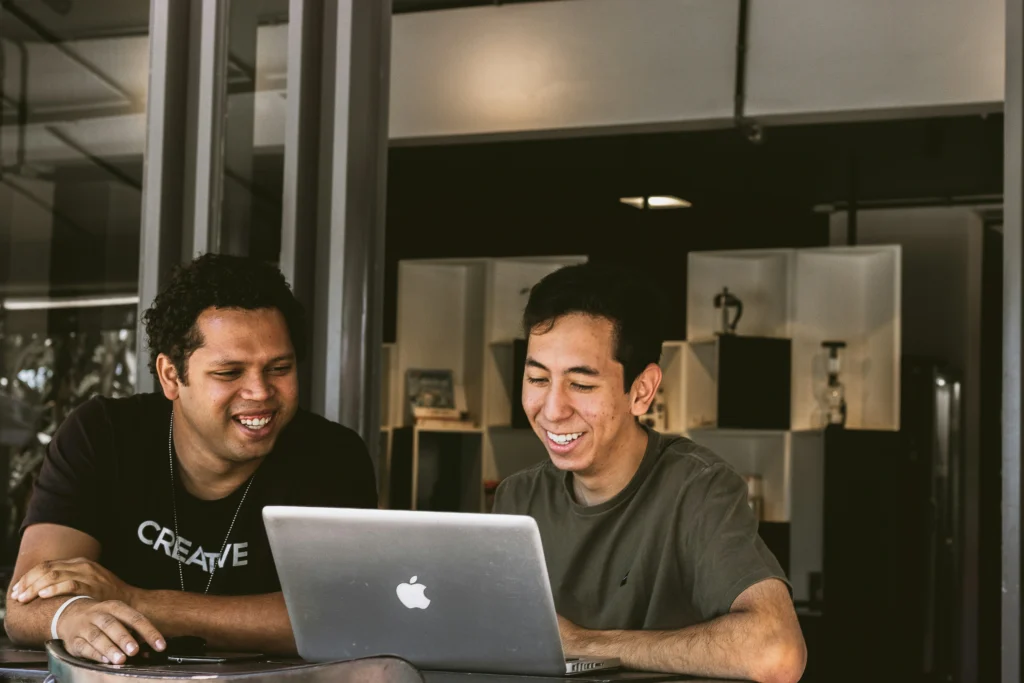 Two young men sit together at a table, smiling warmly as they look at a silver laptop computer screen in a dimly lit indoor setting.