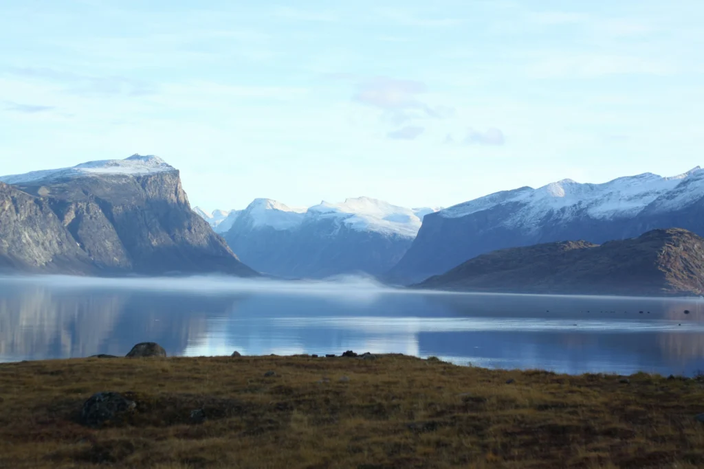 Low fog floats over a calm, mirror-like lake surrounded by dramatic snow-capped mountain peaks and dry grassy tundra under a pale blue sky.