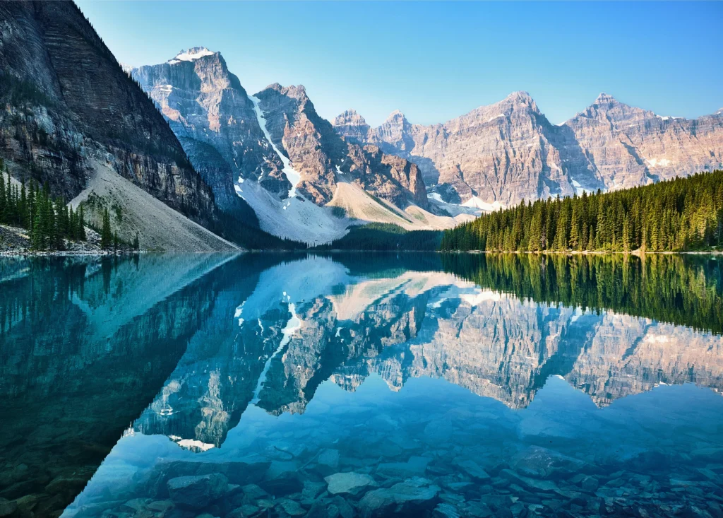 Majestic snow-dusted mountains and green pine trees perfectly reflect in the crystal-clear turquoise waters of Moraine Lake, Canada.