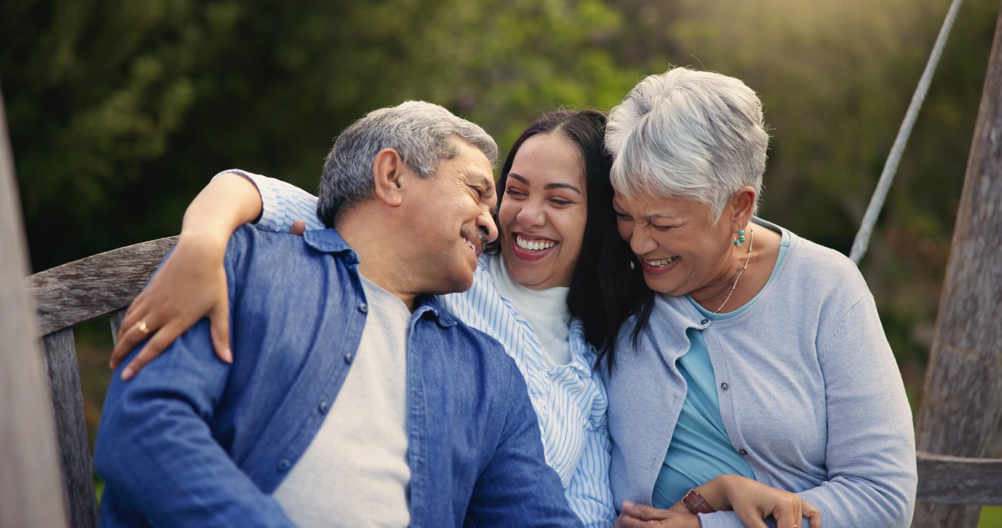 A joyful scene of three people sitting close, smiling brightly. All are wrapped in a warm embrace, conveying happiness and familial love. Greenery is visible behind them.
