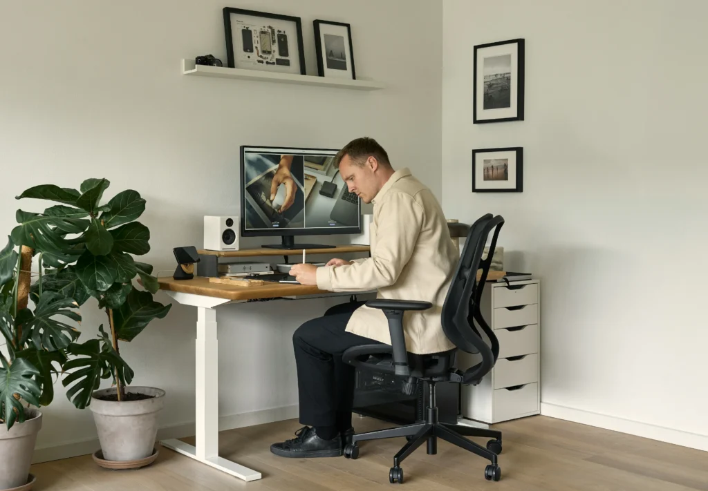 A man sits at a light wood standing desk in a home office, focused on writing with a stylus while looking at a large computer monitor.