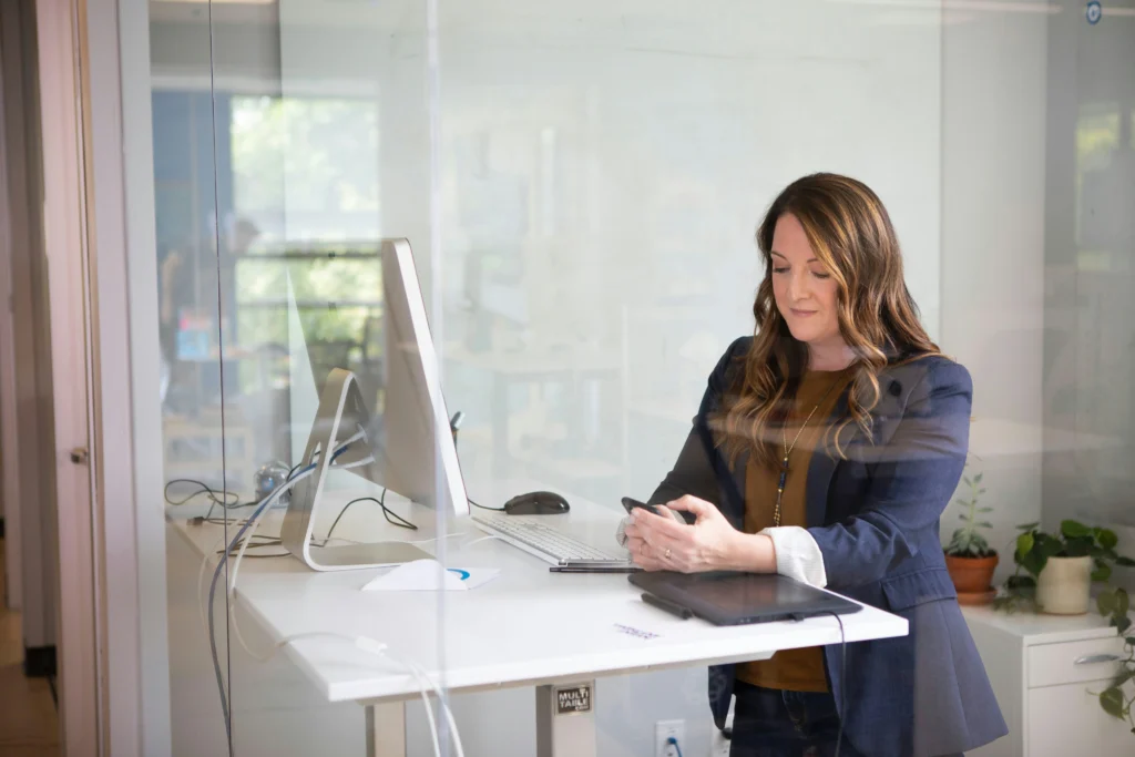 Professional businesswoman wearing a navy blazer stands at a white height-adjustable desk while checking her smartphone in a modern office.