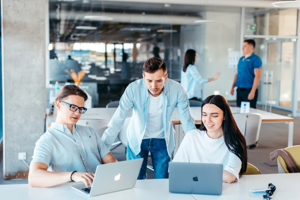 Three young colleagues gather around laptops at a table, discussing work inside a brightly lit, modern open-plan office environment.