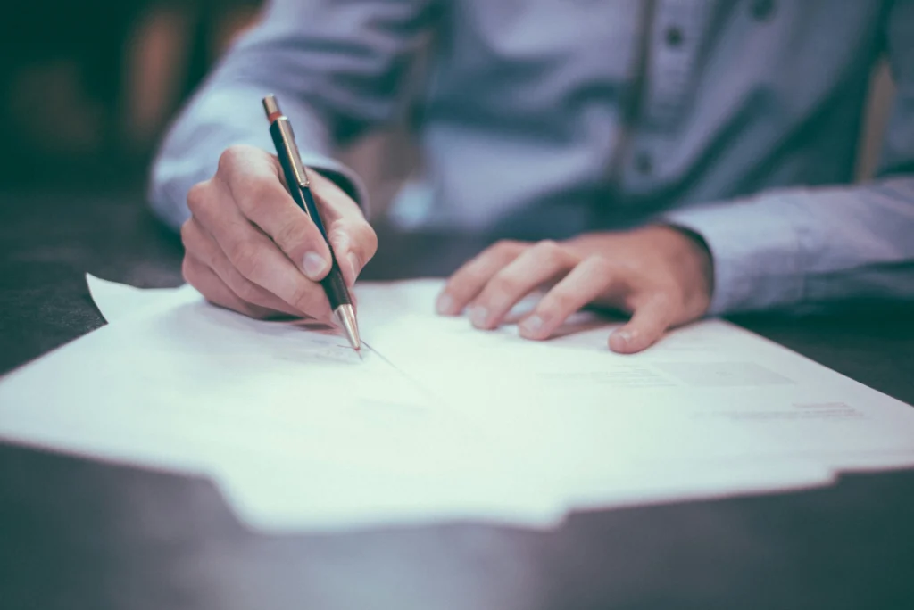 Close-up of a person's hands writing or signing a white paper document with a silver and black pen, wearing a light blue dress shirt.