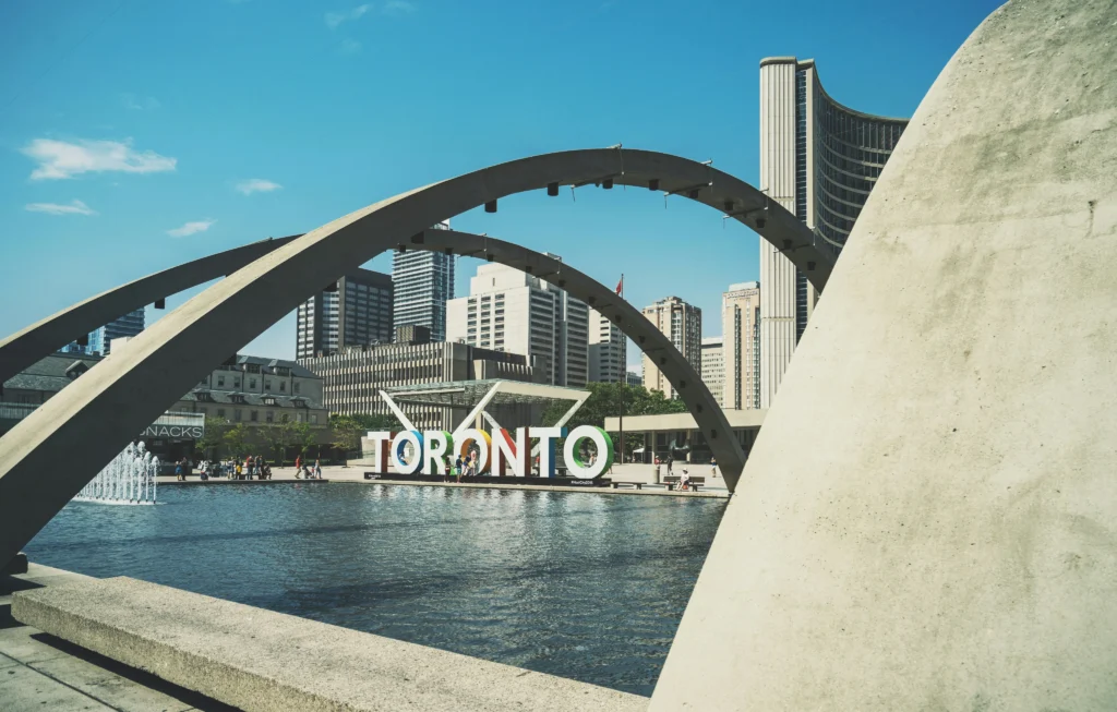 Large 3D Toronto sign sits near a reflecting pool framed by concrete arches, with city skyscrapers and a clear blue sky in the background.