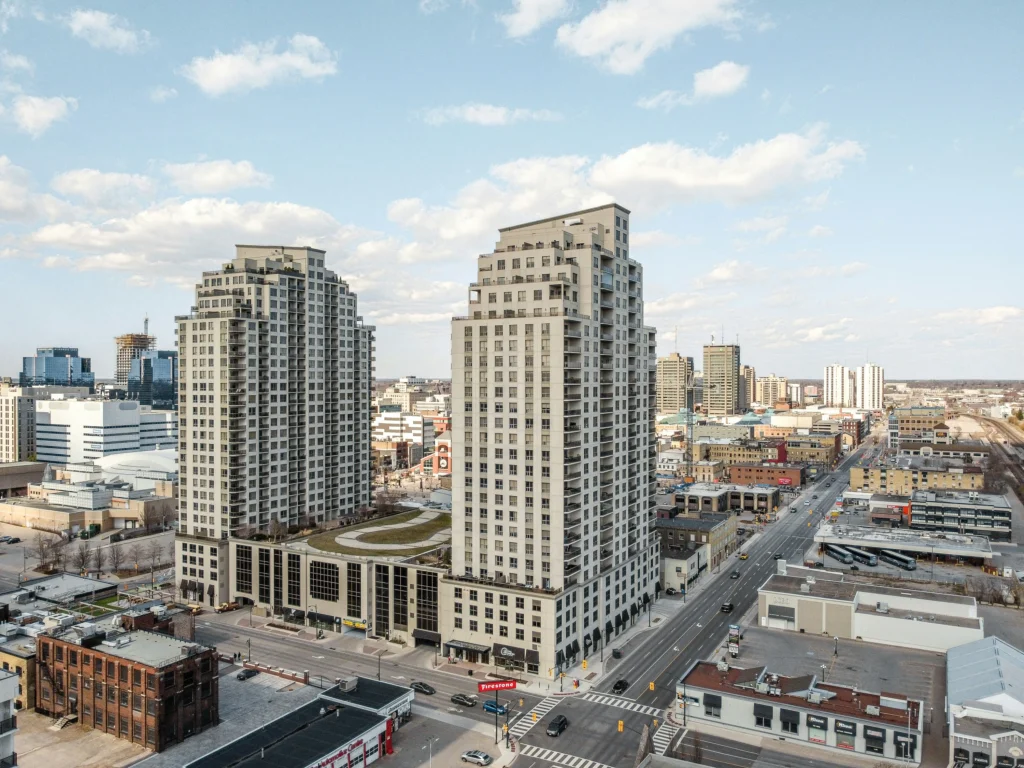 Aerial view of two tall modern residential high-rise buildings situated along a city street lined with lower commercial buildings.