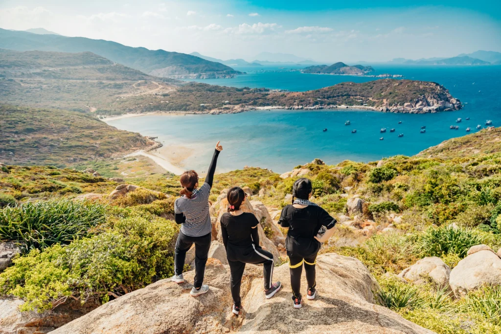 Three hikers stand atop a rocky hill overlooking a scenic coastal bay surrounded by green mountains and blue ocean waters.