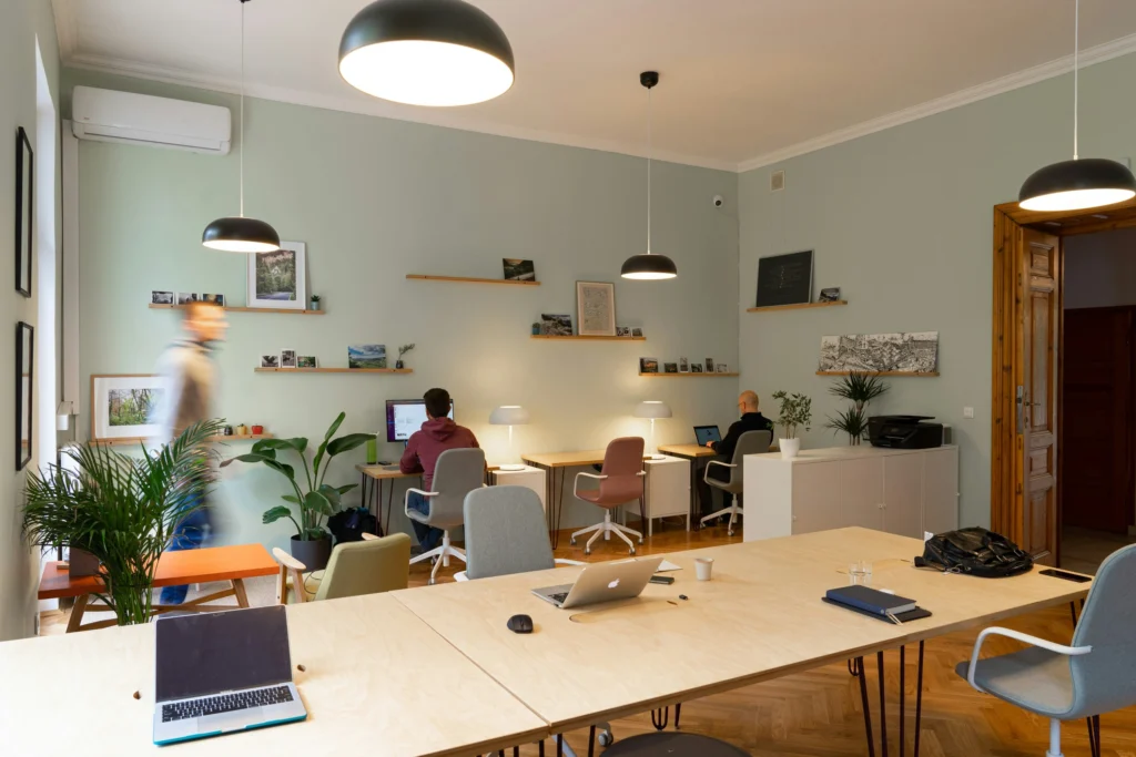 Spacious coworking space featuring individual workstations, warm pendant lighting, indoor plants, and a large communal wooden worktable.