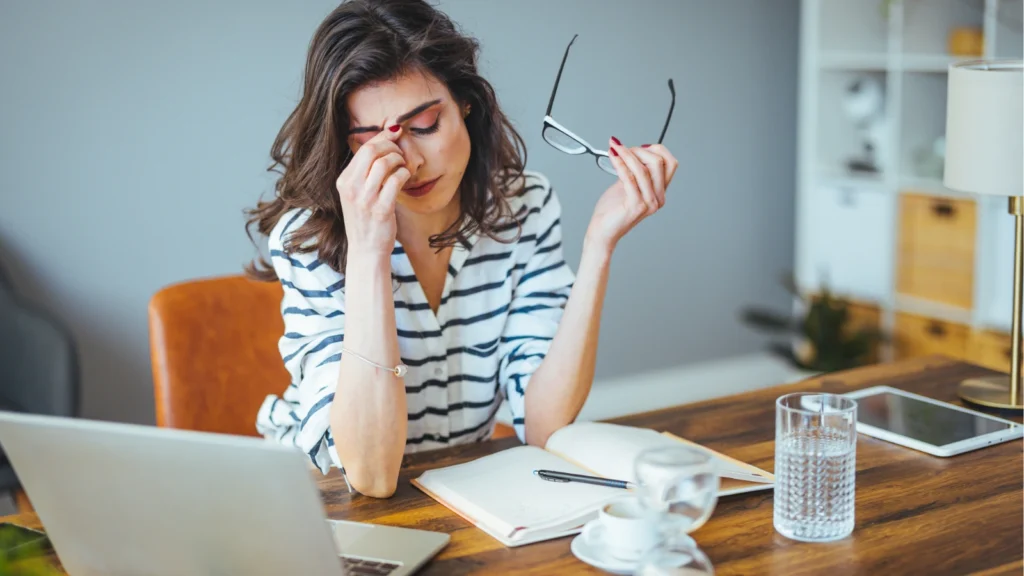 A fatigued woman in a striped shirt sits at a wooden desk, holding her glasses in one hand while pinching the bridge of her nose. A laptop, an open notebook, a glass of water, and a coffee cup are arranged on her workspace, conveying a sense of digital eye strain or exhaustion.