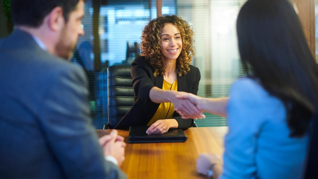A smiling woman with curly hair, wearing a black blazer over a yellow top, reaches across a wooden conference table to shake hands with someone seated opposite her. The back of the person she is shaking hands with, as well as another person sitting beside them in a suit, are visible in the foreground, suggesting a successful job interview or the closing of a business deal.