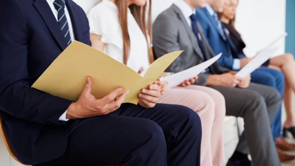 A close-up view of several professionally dressed job candidates sitting in a row in a waiting area. In the foreground, a person in a navy blue suit is reviewing documents inside an open manila folder, while the other candidates sitting beside them hold printed papers.