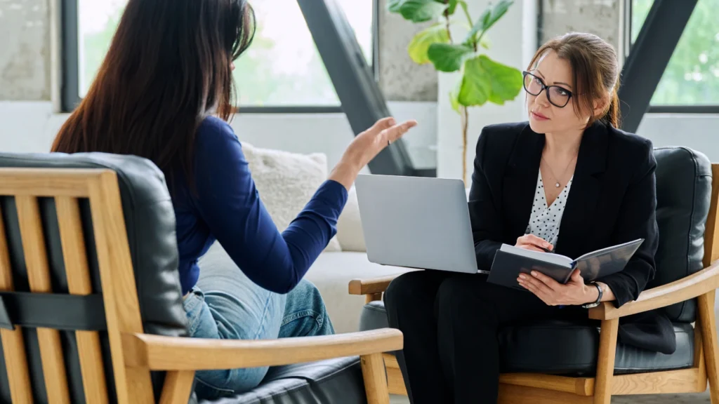 A professional consultation or interview setting where a woman wearing a black blazer and glasses sits in a wood-and-leather chair, balancing a laptop on her lap while taking notes in a notebook. She is listening attentively to a woman in a blue long-sleeved shirt and jeans who is actively gesturing with her hand.