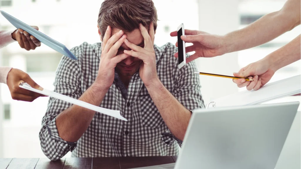 A stressed man in a checkered shirt sits at a desk with his face buried in his hands. Multiple hands from off-screen surround him, aggressively thrusting a smartphone, a tablet, paperwork, and a pencil into his space.