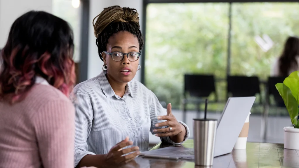 A woman with braided hair and glasses gestures dynamically while speaking across a desk with a laptop during a meeting.