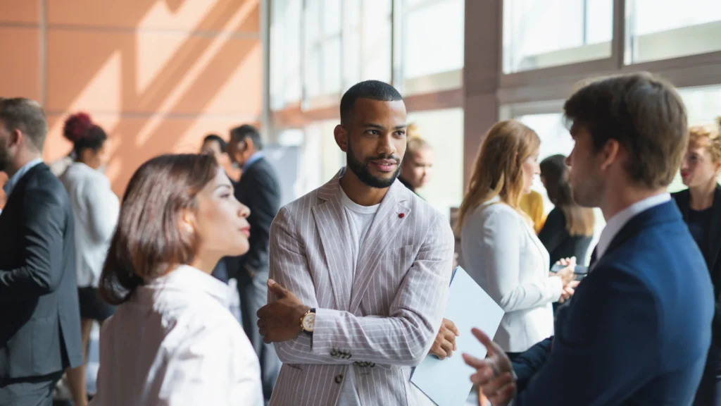 A stylishly dressed man in a pinstriped blazer listens attentively while networking with colleagues in a sunlit atrium.