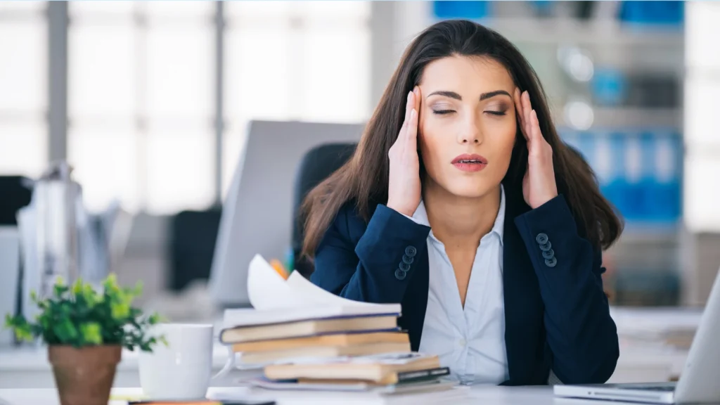 Stressed businesswoman in a dark blue blazer sitting at her office desk with her eyes closed and hands resting on her temples next to stacks of paper.