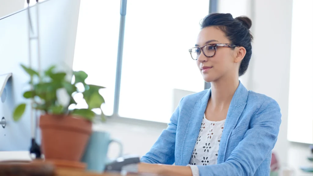 A woman with glasses and a blue blazer sitting at a desk and smiling at a computer screen next to a potted plant.