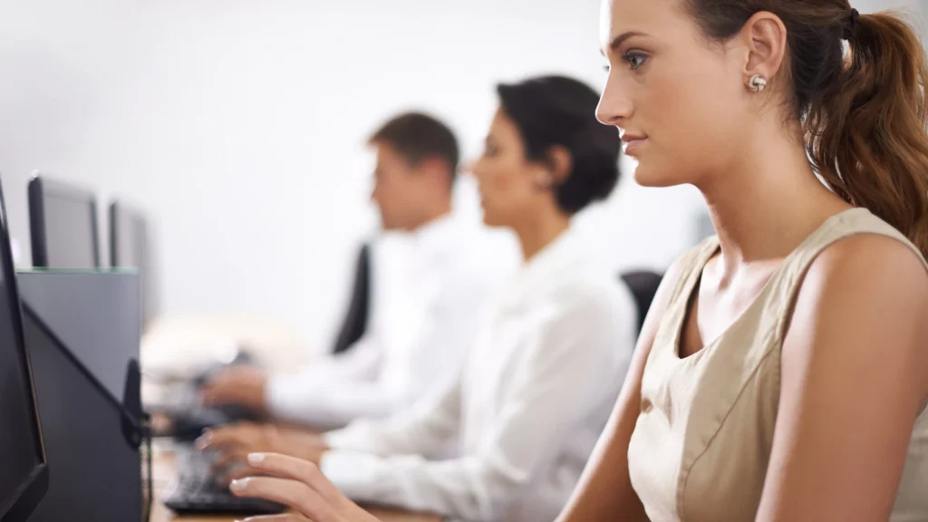 Side view of a woman wearing a beige top typing on a computer keyboard in an office setting alongside colleagues.