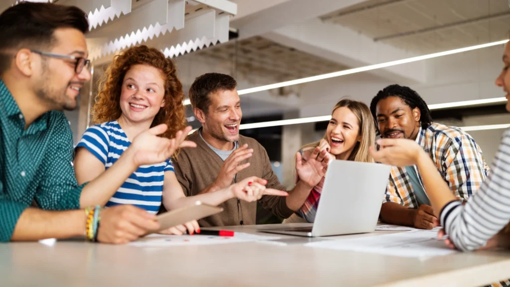 A team of diverse colleagues sit around a conference table laughing and gesturing enthusiastically during a collaborative laptop meeting.