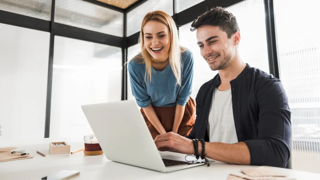 A smiling man and woman look at a laptop screen together while collaborating on a project at a white desk in a modern glass office space.