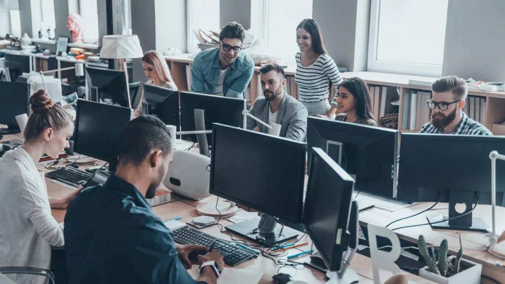 A busy office scene filled with creative professionals sitting at shared wooden desks and collaborating in front of desktop computer monitors.