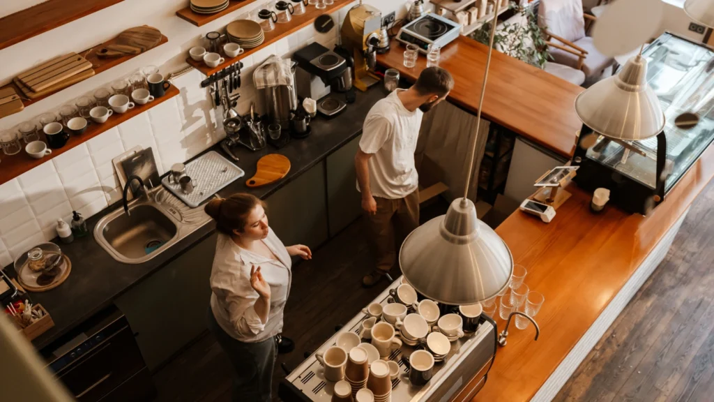 A high-angle top-down view of two cafe baristas working behind a long wooden counter surrounded by coffee cups and espresso equipment.