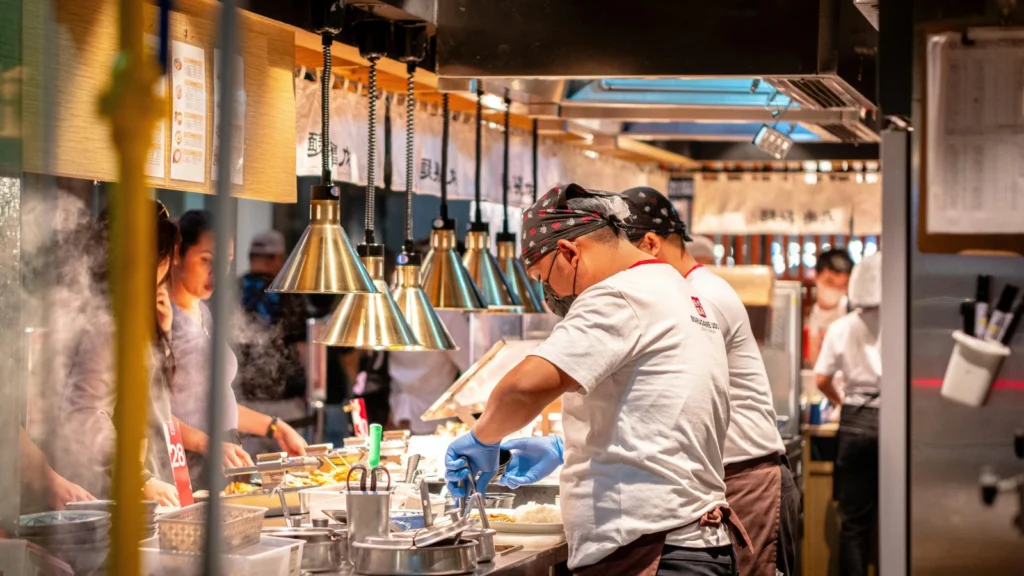 Kitchen staff wearing blue gloves, aprons, and bandana head coverings preparing food behind a glass counter under hanging heat lamps in a busy restaurant.
