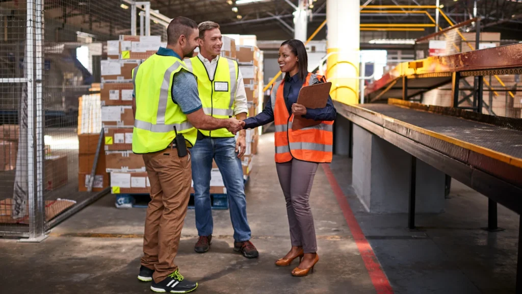 A female manager in a high-visibility orange vest holding a clipboard and shaking hands with a male logistics worker in a yellow safety vest inside a warehouse, while another colleague looks on.