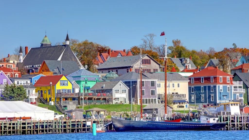 A scenic waterfront view of Lunenburg, Nova Scotia, featuring colorful coastal buildings, historical wooden architecture, and a large black sailboat docked at the pier on a clear day.