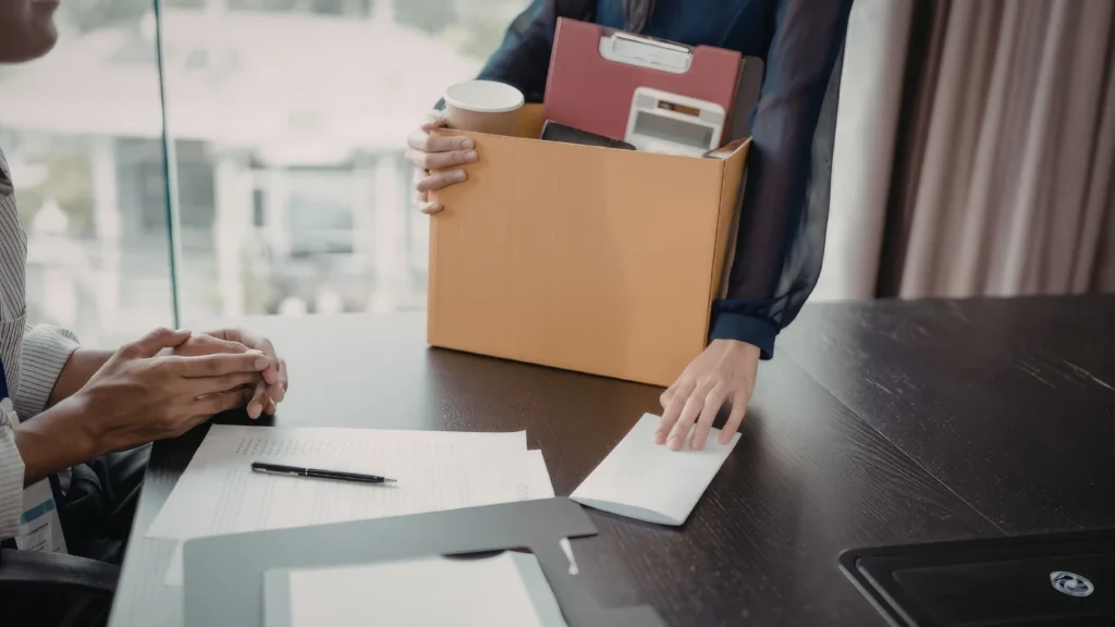 An employee holding a cardboard box filled with office items, such as folders and a calculator, placing a white envelope onto a desk in front of a seated colleague whose hands are clasped together.