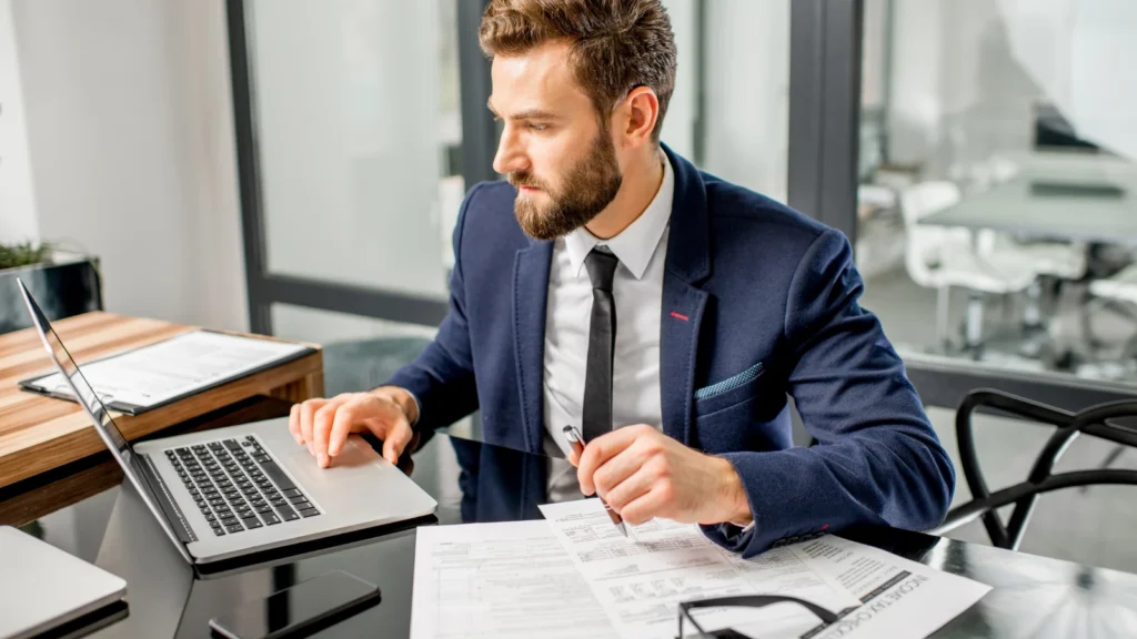 A bearded businessman in a dark blue suit and tie sitting at a black glass desk, looking at a laptop screen while holding a pen over a printed document.