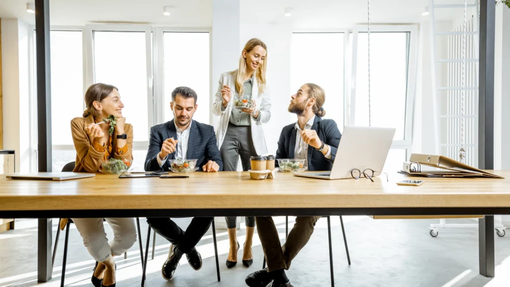 Four business professionals sit together around a large wooden table inside a bright open office space, enjoying a healthy lunch break.