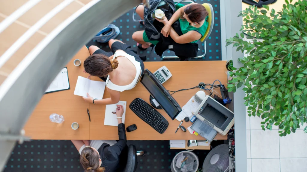 A high-angle overhead view of two office employees reviewing paperwork together at a large wooden desk equipped with a computer.