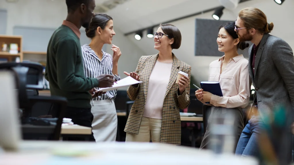 A group of five diverse corporate professionals stand together in an open-plan office space, chatting casually while holding notebooks.