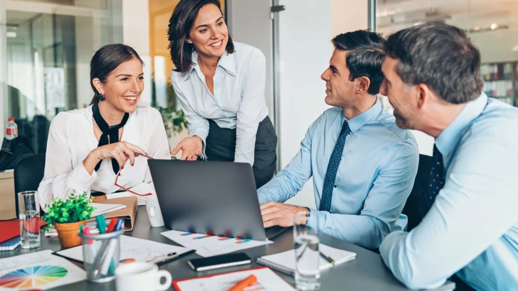 Four business professionals smile and engage in discussion around a conference table while looking at reports on an open laptop screen.