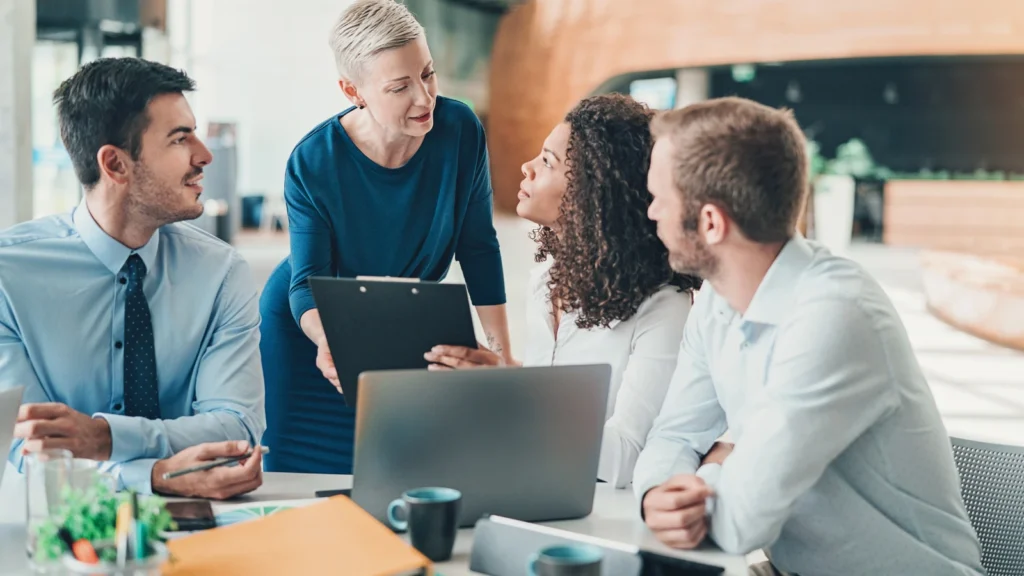 A female manager holds a clipboard while leaning over a table to review documents during a team meeting with three seated colleagues.