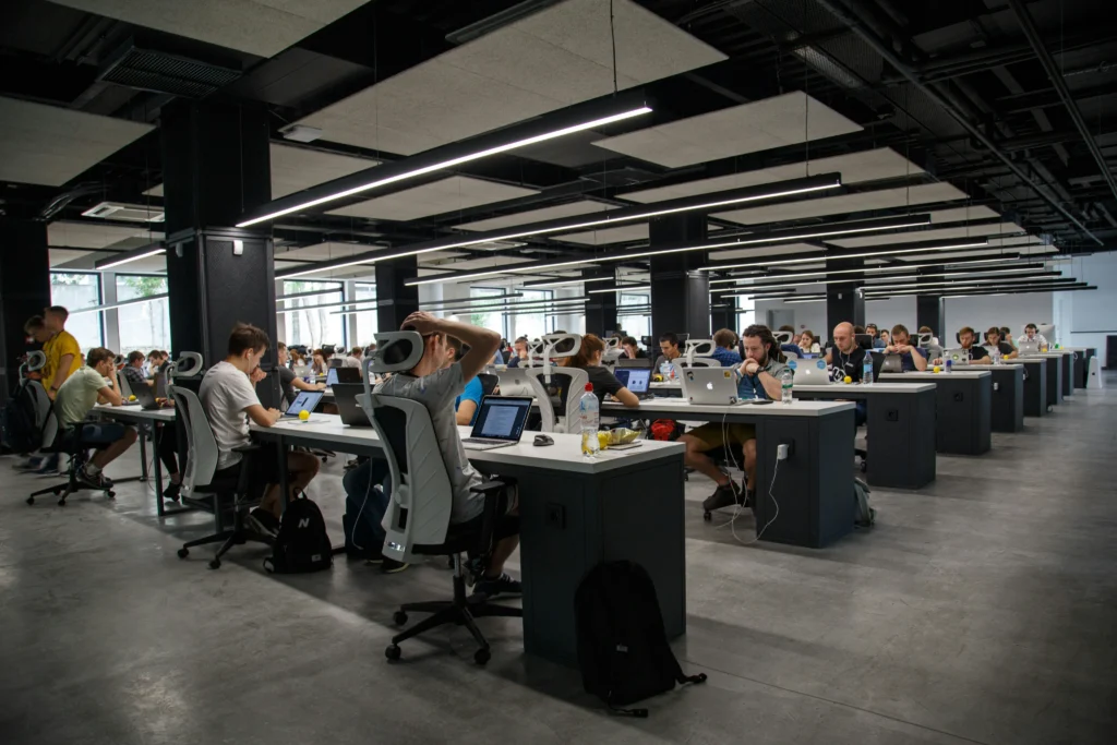 Dozens of tech workers sit at long rows of white desks working on laptops inside a large, open-plan office under linear overhead lights.